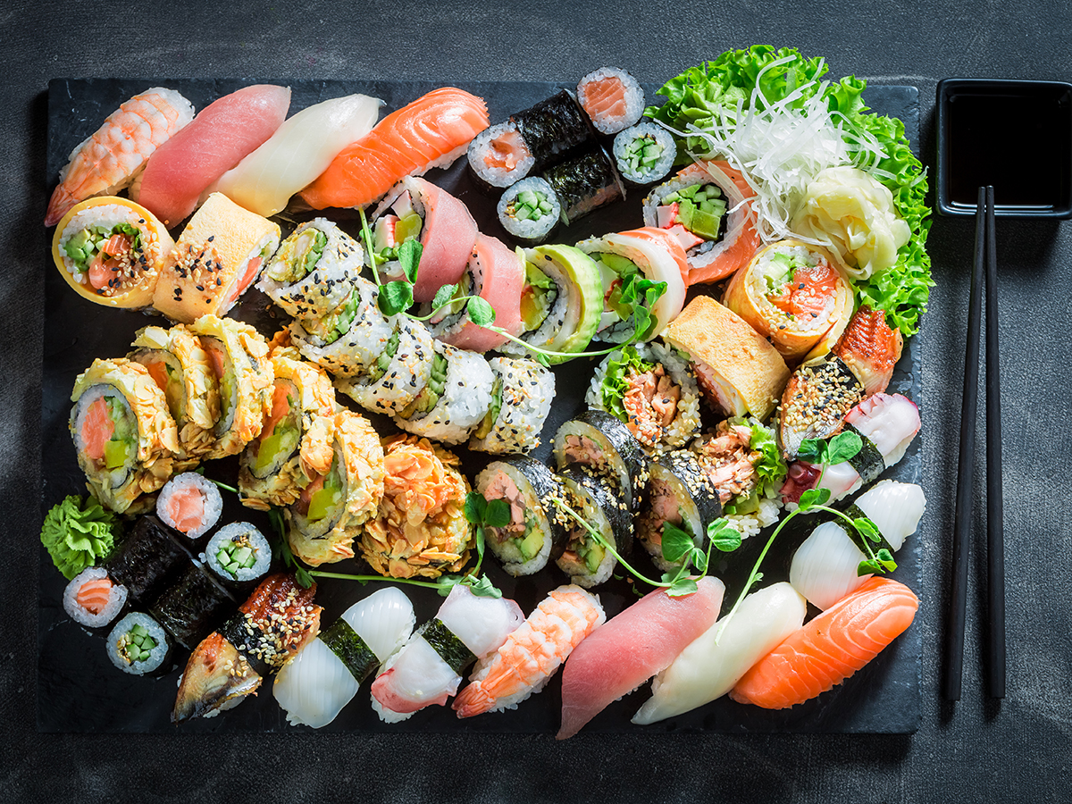 Top‑view large assorted sushi platter on dark slate, includes nigiri, many maki rolls, pickled vegetables, lettuce garnish, black chopsticks and small soy‑sauce dish on side.