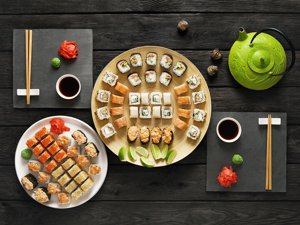 Flat‑lay shot of assorted sushi spread on dark wood table, round bamboo platter, white plate, green teapot, lime wedges, ginger, wasabi, soy sauce bowls and wooden chopsticks.