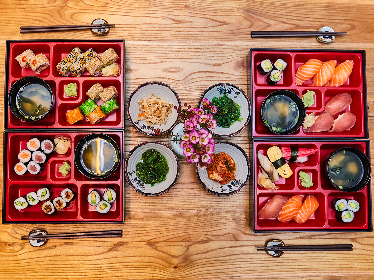Overhead view of four red Japanese bento boxes filled with sushi, maki and miso soup, side small bowls of seaweed salad and kimchi, tiny flower decor on light wooden table.