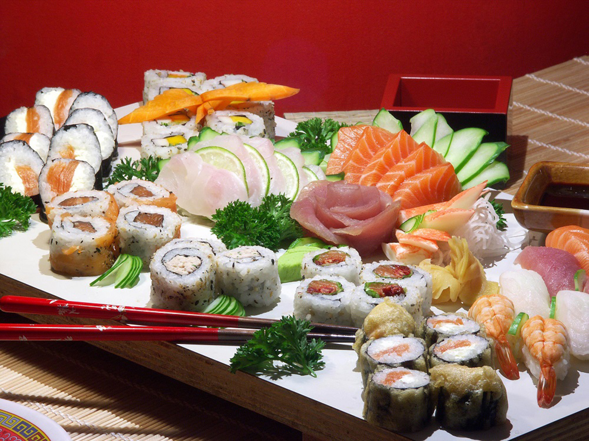 Large mixed sushi and sashimi platter, diverse maki rolls, fresh salmon and white fish sashimi, garnished with parsley and cucumber slices, red chopsticks rest on the plate against red wall background.