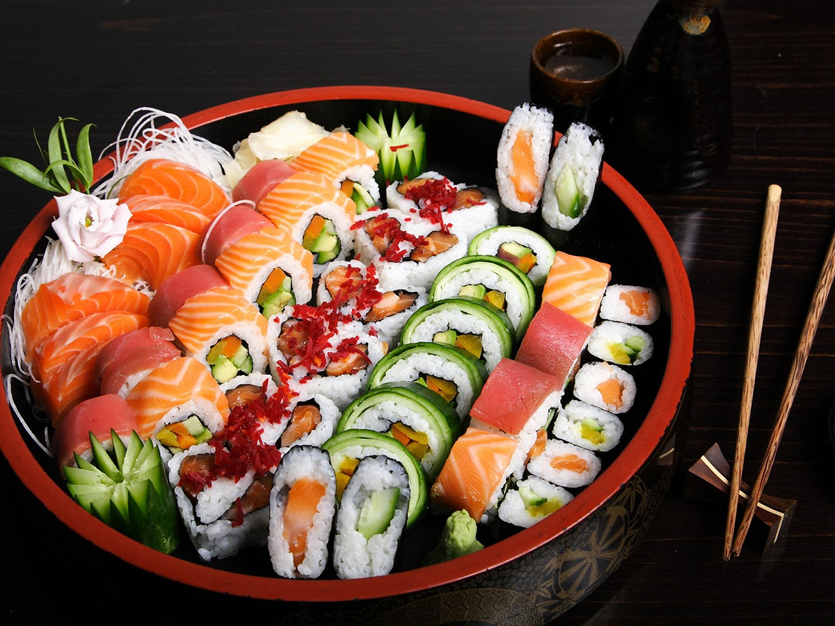 Round lacquered sushi platter filled with salmon, tuna nigiri and varied maki rolls, decorated with carved cucumber, flower garnish, wasabi, wooden chopsticks on dark wooden table.