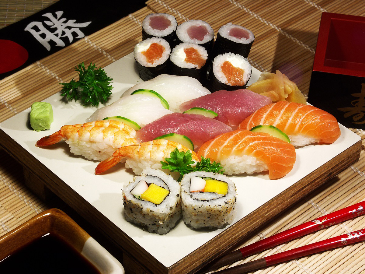 Assorted sushi platter on white wooden board, contains nigiri with shrimp, salmon, tuna, maki rolls, wasabi and garnish, set on bamboo mat with red chopsticks and soy‑sauce dish nearby.