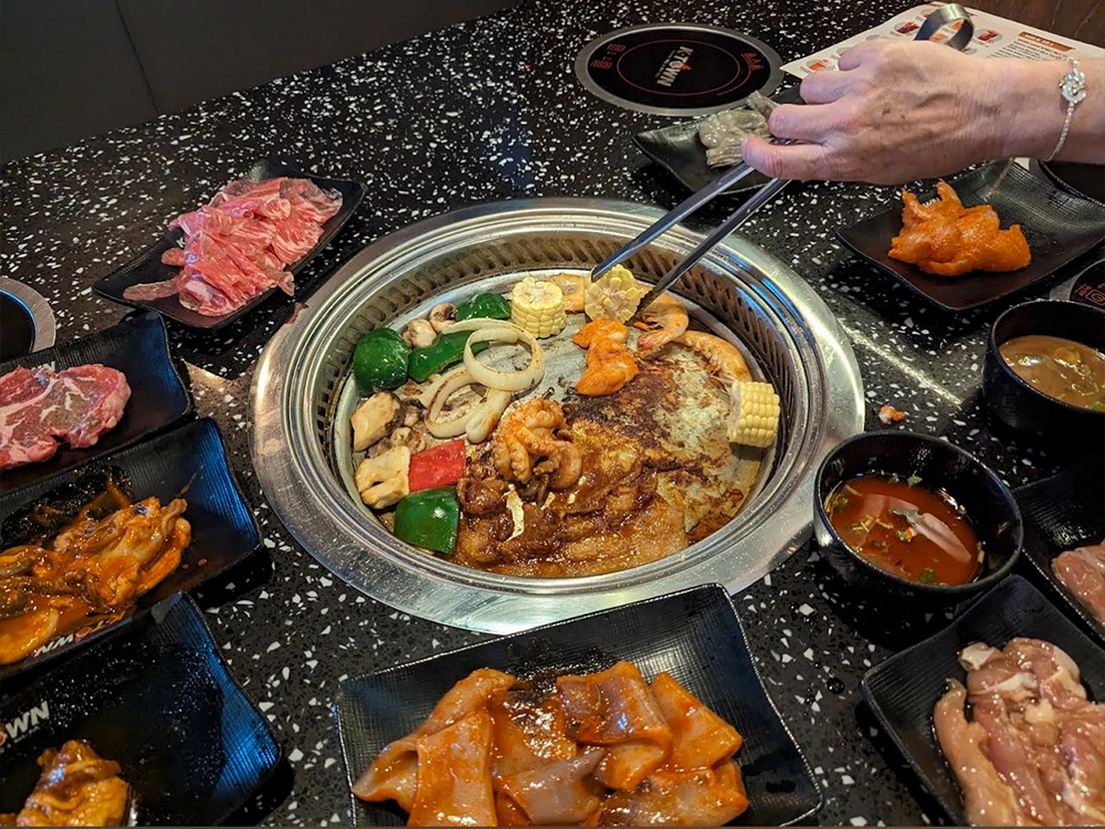 The dining table is rich in ingredients, including raw meat and side dishes, with a baking tray in the middle at K-Town Korean BBQ & Hotpot, a Korean BBQ & Hotpot Restaurant in Fort Wayne