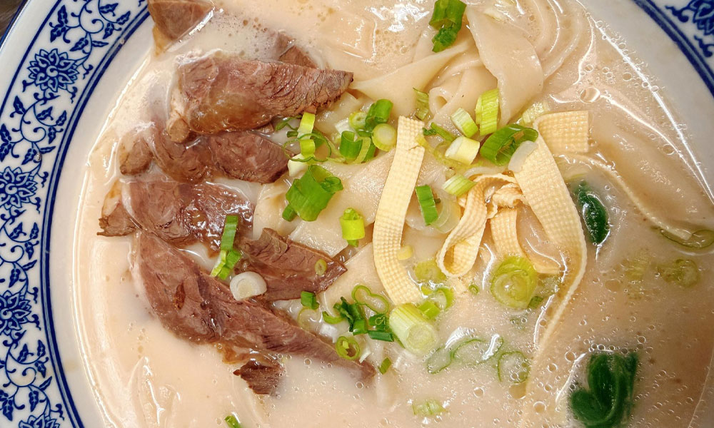 Creamy beef wide noodle soup with sliced beef, tofu strips and chopped green onions in blue floral ceramic bowl at Kung Fu Noodle House, a Chinese Restaurant in West Allis