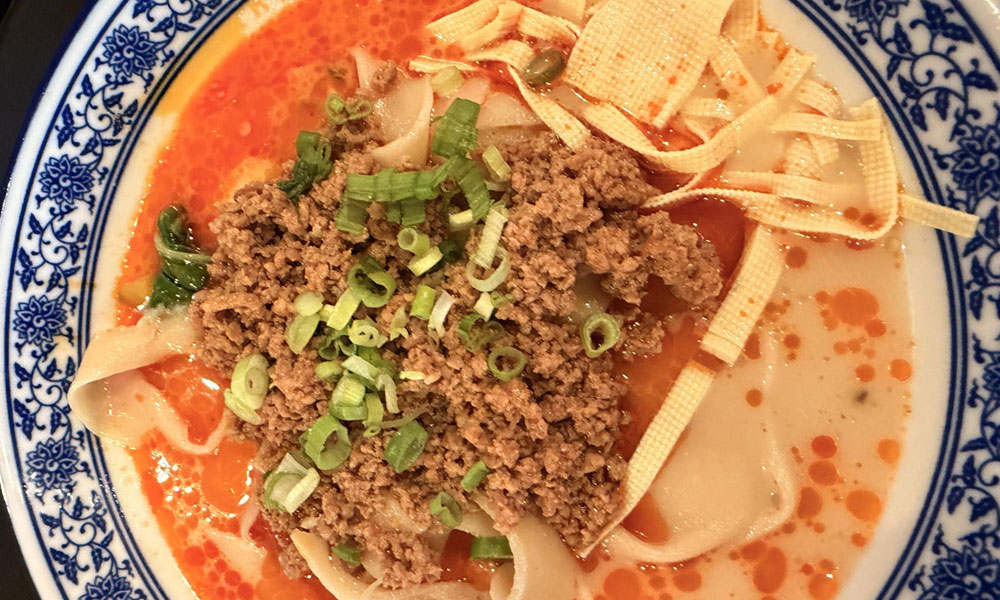 Spicy minced pork wide noodle soup with red chili oil, tofu skin and fresh green onion garnish on patterned porcelain plate at Kung Fu Noodle House, a Chinese Restaurant in West Allis