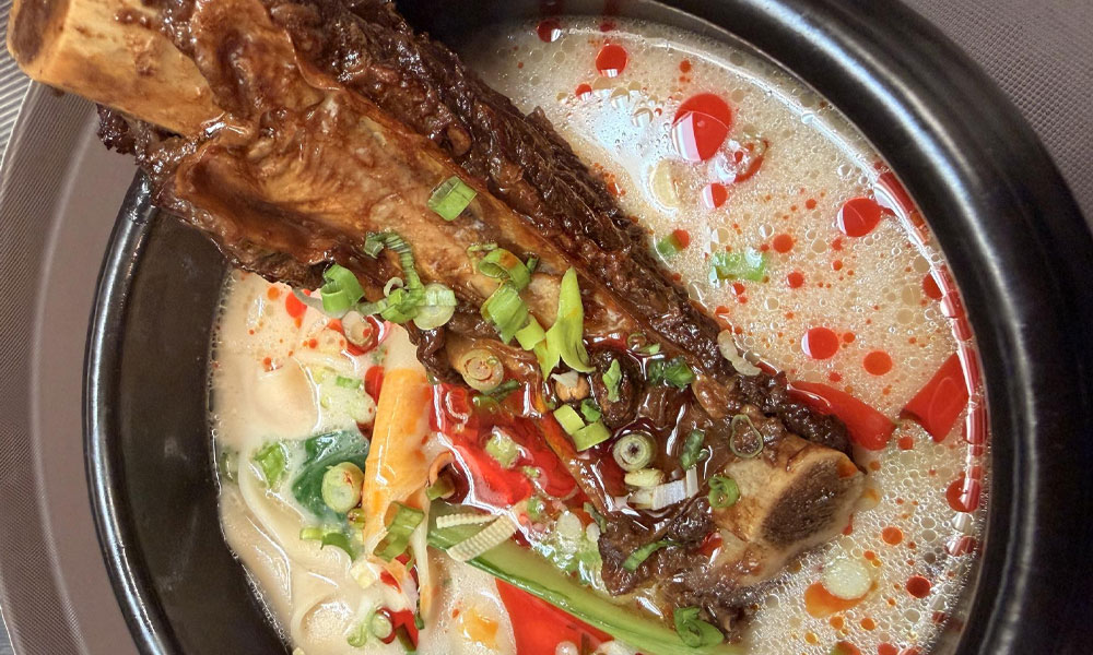 Braised beef rib bone noodle soup in black metal pot with creamy broth, chili oil, veggies and sliced scallions at Kung Fu Noodle House, a Chinese Restaurant in West Allis