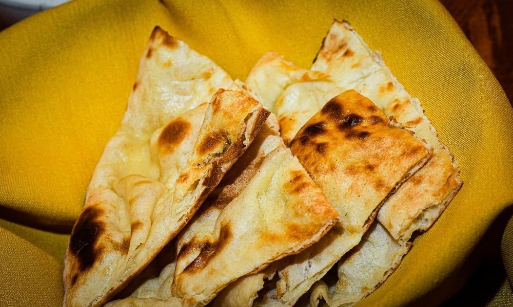 Close-up of freshly baked triangular naan bread slices with golden-brown charred marks. Stacked and served in a basket lined with soft bright yellow fabric.