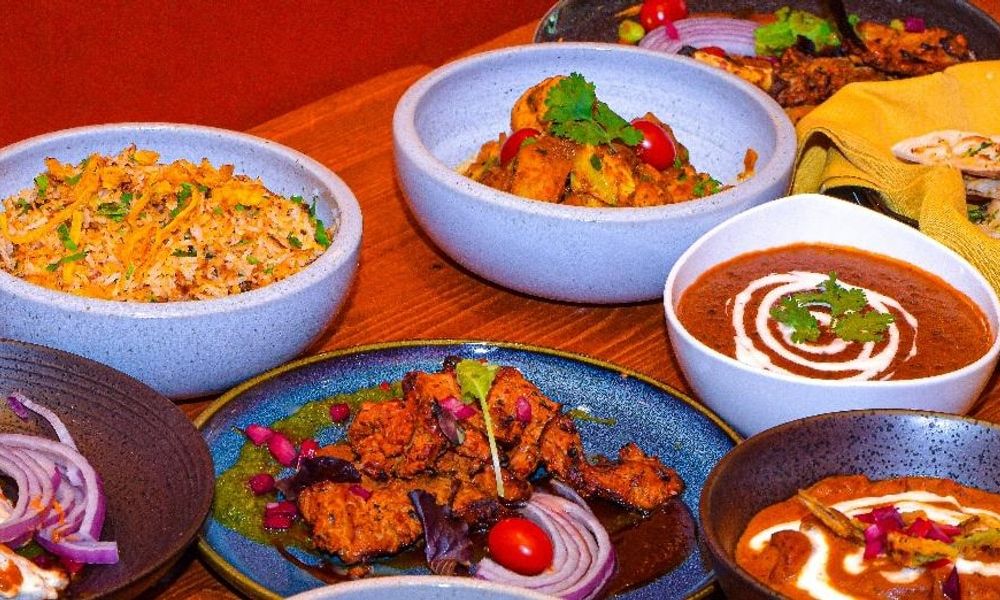 Tabletop view of multiple bowls and plates filled with Indian feast, including biryani rice, butter chicken, grilled tandoori meat, lentil curry, garnished with cilantro, cherry tomatoes and sliced red onion.