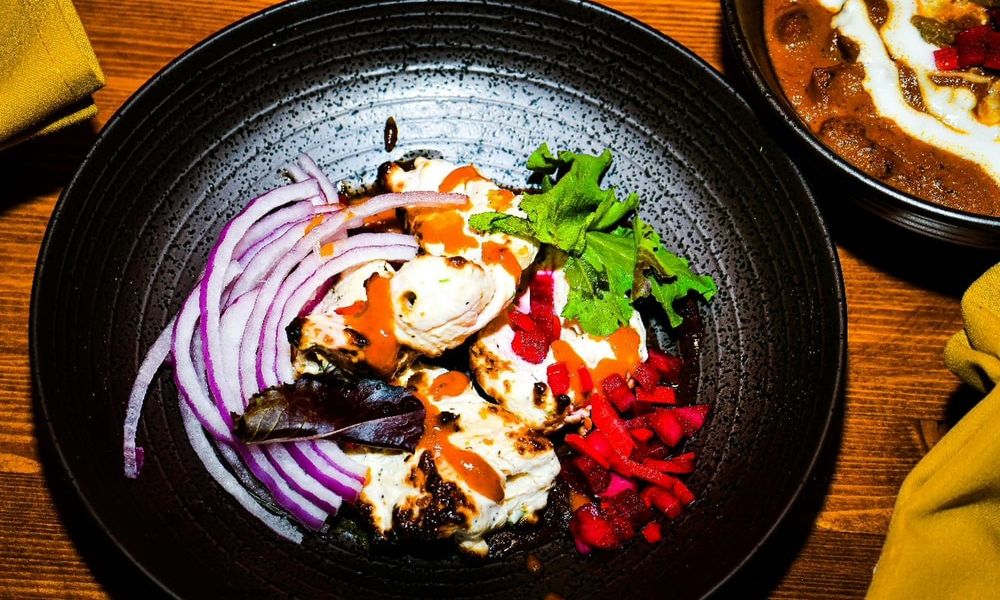 Top-down view of grilled chicken tikka on dark speckled plate. Sliced purple onion, fresh leaf lettuce, diced red pickled vegetables and orange sauce drizzle accompany the tender charred chicken pieces.