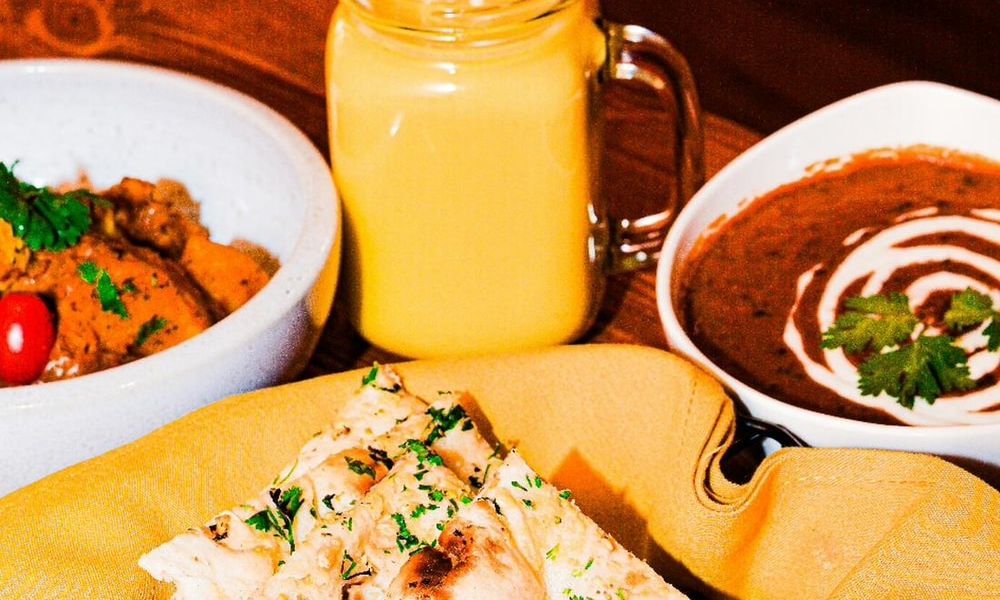 Close-up of Indian food on wooden table, two bowls of creamy curry garnished with cilantro, fresh garlic naan wrapped in yellow cloth, and glass mason jar filled with bright golden mango lassi.
