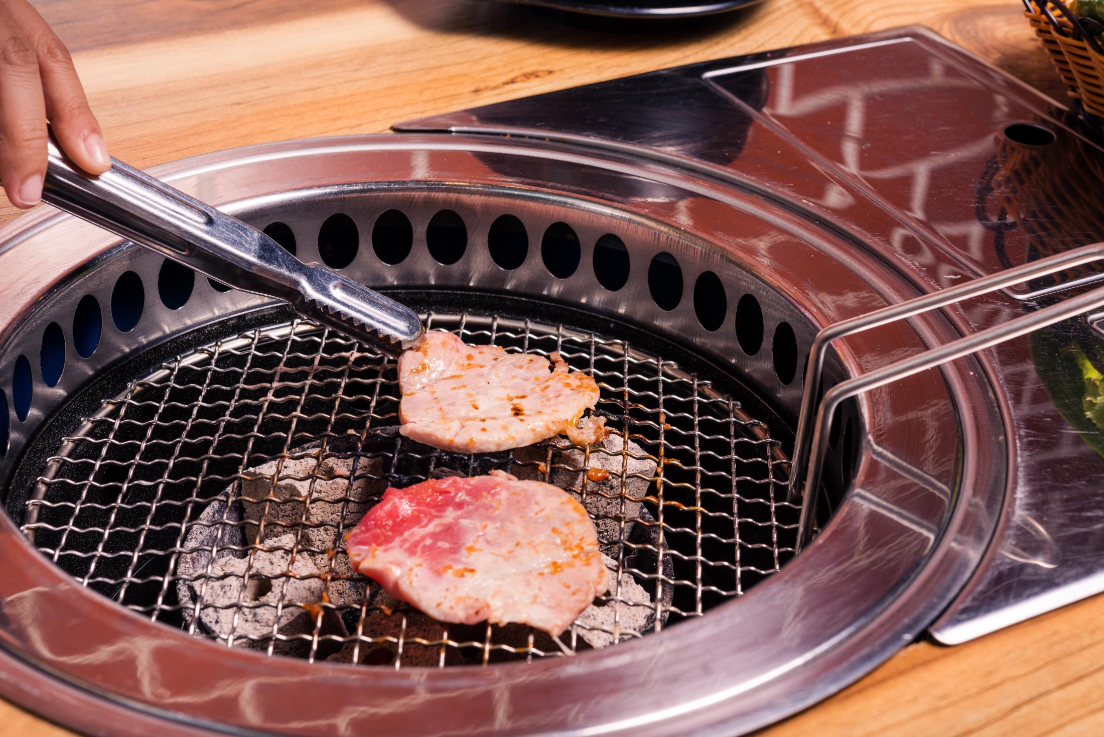 Hand using tongs to grill marinated pork slices on a Korean BBQ charcoal grill at Legend Pot, Hot Pot Restaurant in Baton Rouge