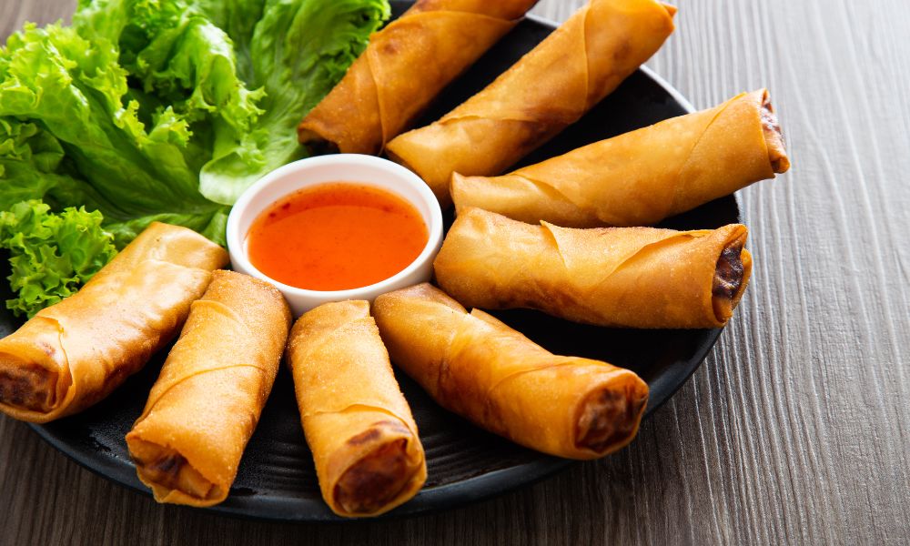 Crispy fried spring rolls arranged on black plate, served with sweet chili dipping sauce and fresh lettuce leaves on wooden table.