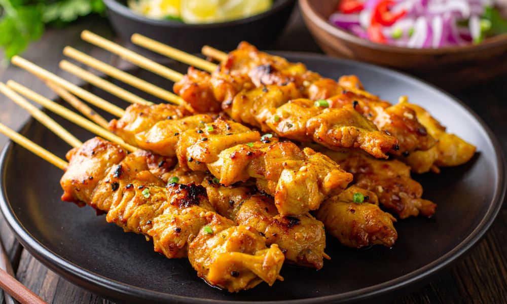 Grilled marinated chicken satay skewers on black plate, garnished with green scallions, side bowls of chopped vegetables in blurred background.