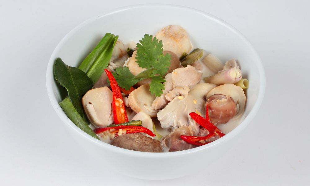 Thai coconut chicken soup in white bowl, filled with chicken pieces, mushrooms, red chili slices, cilantro, lemongrass and kaffir lime leaves.