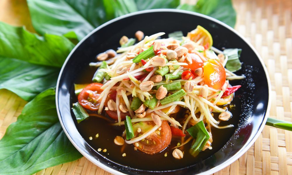 Spicy green papaya salad on black plate, mixed with tomato, long beans, peanuts, chili, savory dressing, set over woven rattan mat with green leaves.