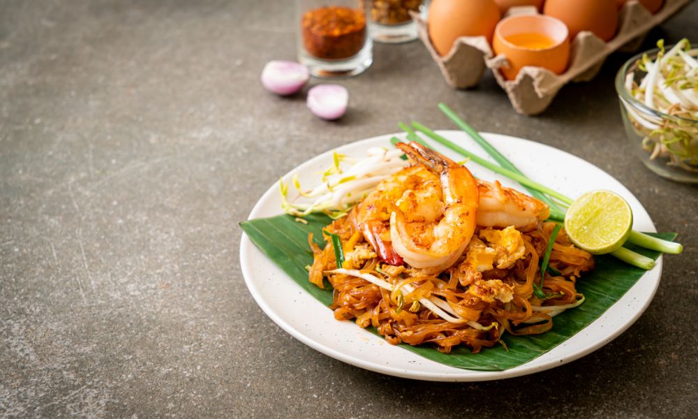 Pad thai stir‑fried rice noodles on white plate over banana leaf, topped with large shrimp, bean sprouts, lime wedge, raw ingredients on concrete background.