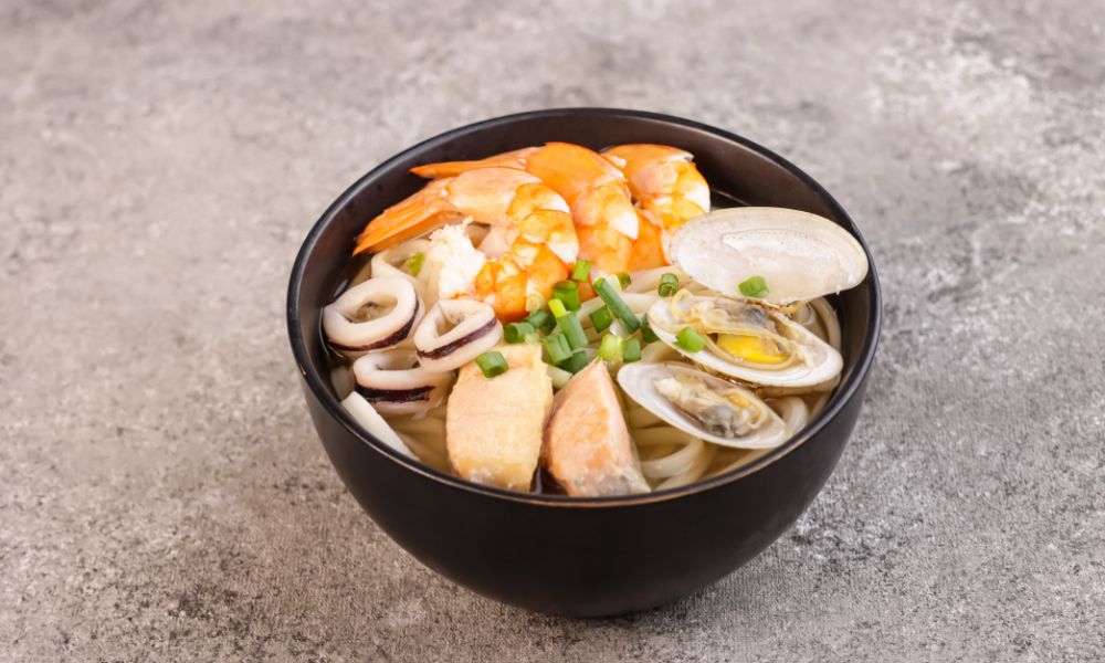 Seafood udon noodle soup in black bowl, loaded with shrimp, clams, squid rings, fish tofu and chopped green onions on grey textured countertop.