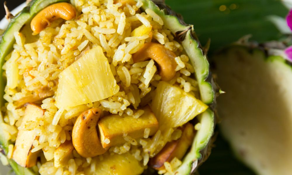 Top‑down close‑up of pineapple fried rice served inside hollowed pineapple shell, mixed with cashew nuts, pineapple chunks and tofu pieces.
