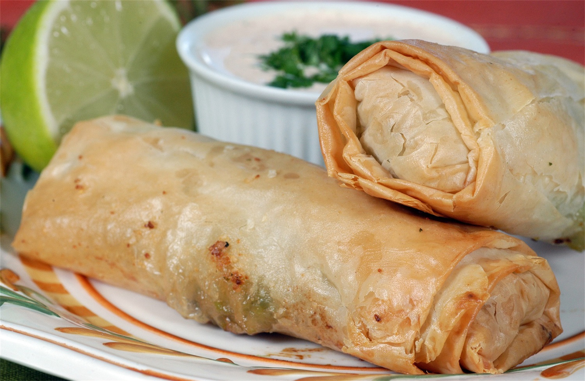 Two crispy spring rolls on a plate, one cut open showing a flaky interior, with lime wedge and dipping sauce.