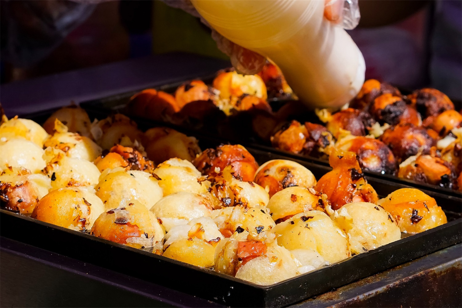 Close-up of takoyaki balls in a black tray, with a hand squeezing mayonnaise on top