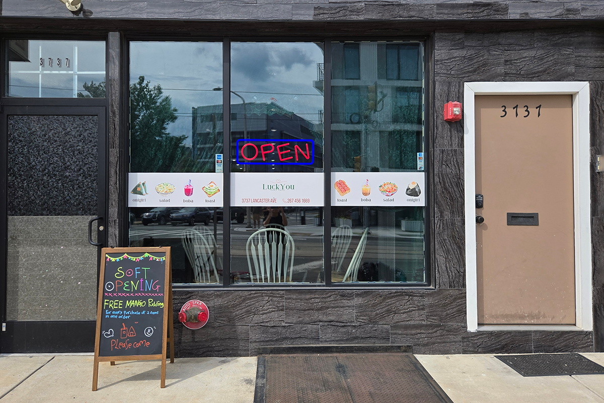 Luckyou-Cozy-Cafe-Asian-Fusion-Café-Exterior-Philadelphia Exterior of LuckYou Cozy Cafe, with dark gray stone wall, glass door and window, “OPEN” sign and food images on the window, accompanied by a opening - promotion chalkboard