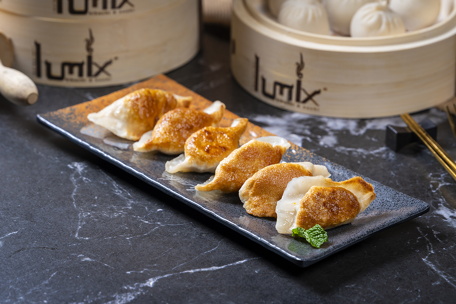 Six crispy pan-fried dumplings on dark speckled rectangular plate. Black marble tabletop, bamboo steamers with buns in background, garnished with mint leaf.