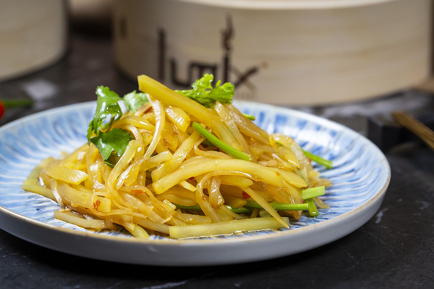 Cold shredded potato salad garnished with cilantro and green onion on blue patterned plate. Black marble tabletop with bamboo steamers in blurred background.