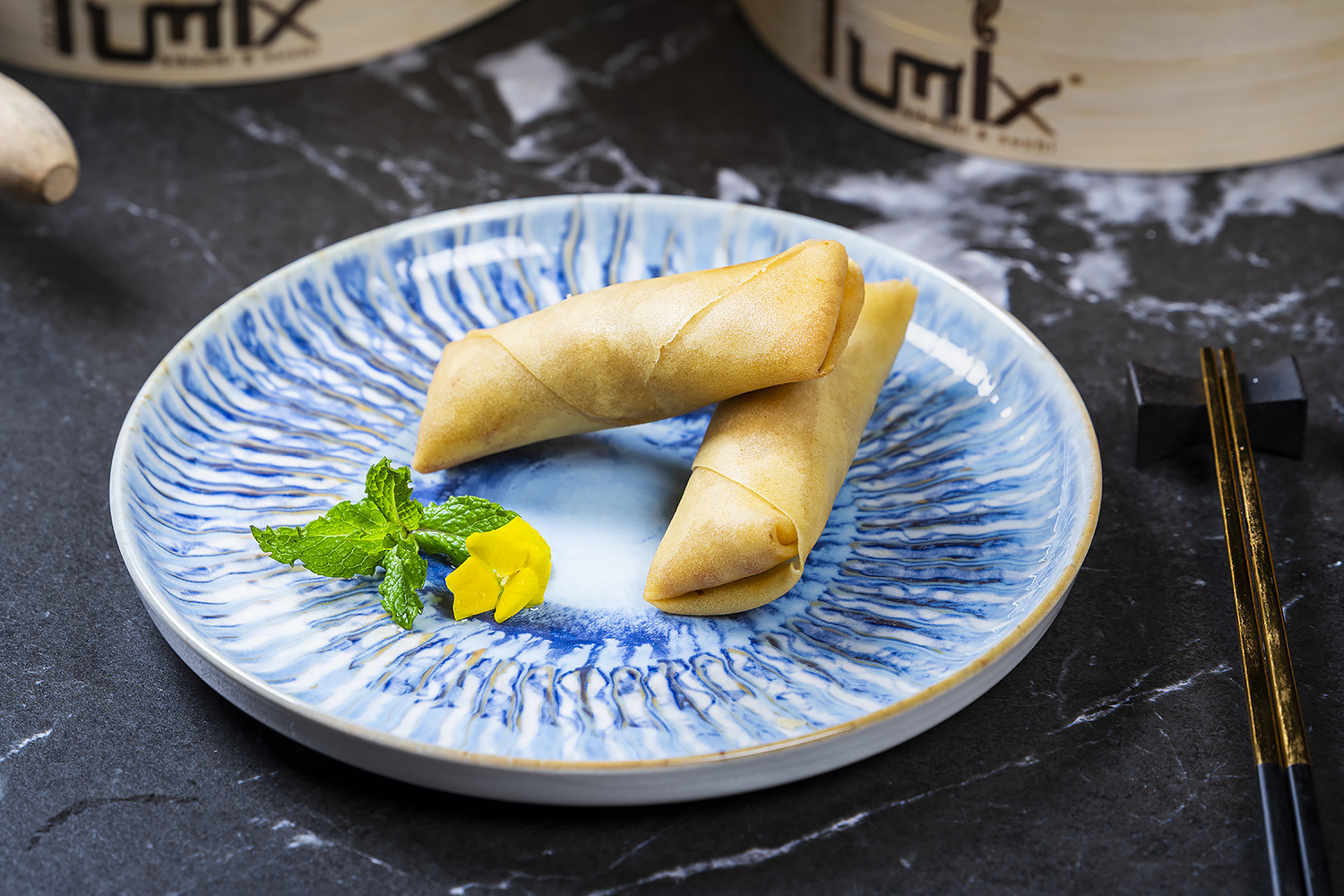 Two golden fried spring rolls stacked on blue patterned ceramic plate. Garnished with mint and edible flower, black marble table with chopsticks beside.