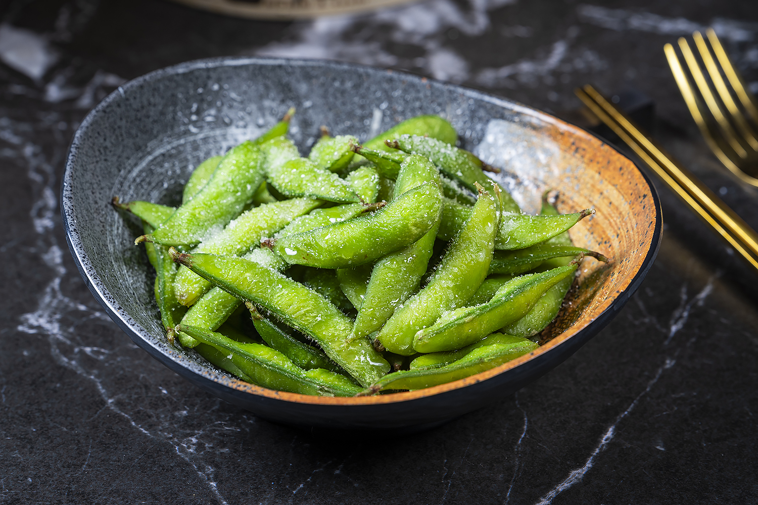 Salted green edamame sprinkled with coarse salt, served in dark speckled bowl. Black marble tabletop, golden fork and chopsticks on the right side.