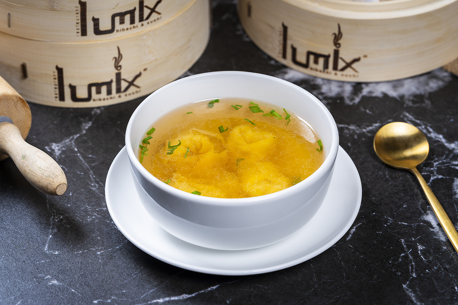 Wonton soup topped with chopped green onions in white bowl on matching saucer. Black marble table, bamboo steamers and golden spoon in background.