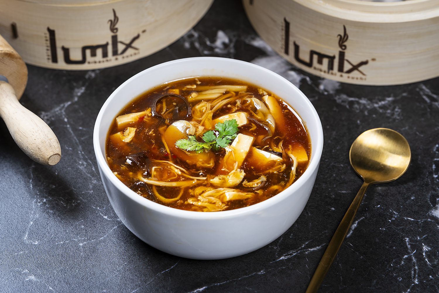 Rich brown hot and sour soup with tofu, wood ear mushrooms, egg strips and cilantro. Black marble table, bamboo steamers and golden spoon beside.