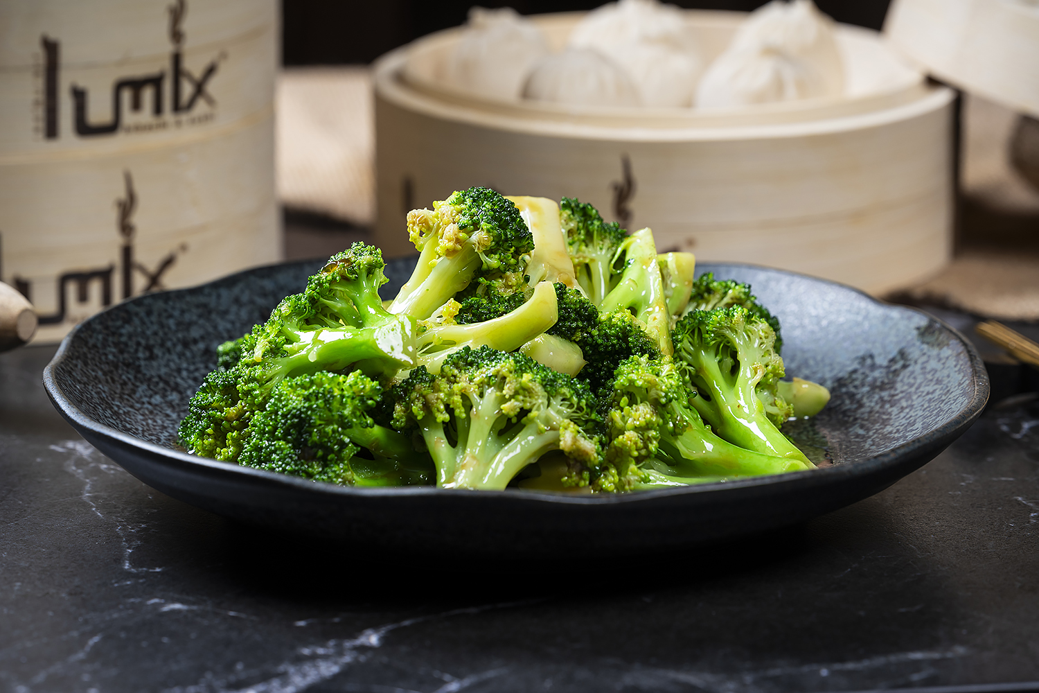 Glazed seasoned broccoli florets on dark textured plate. Bamboo steamers with soup dumplings in blurred background, black marble tabletop.