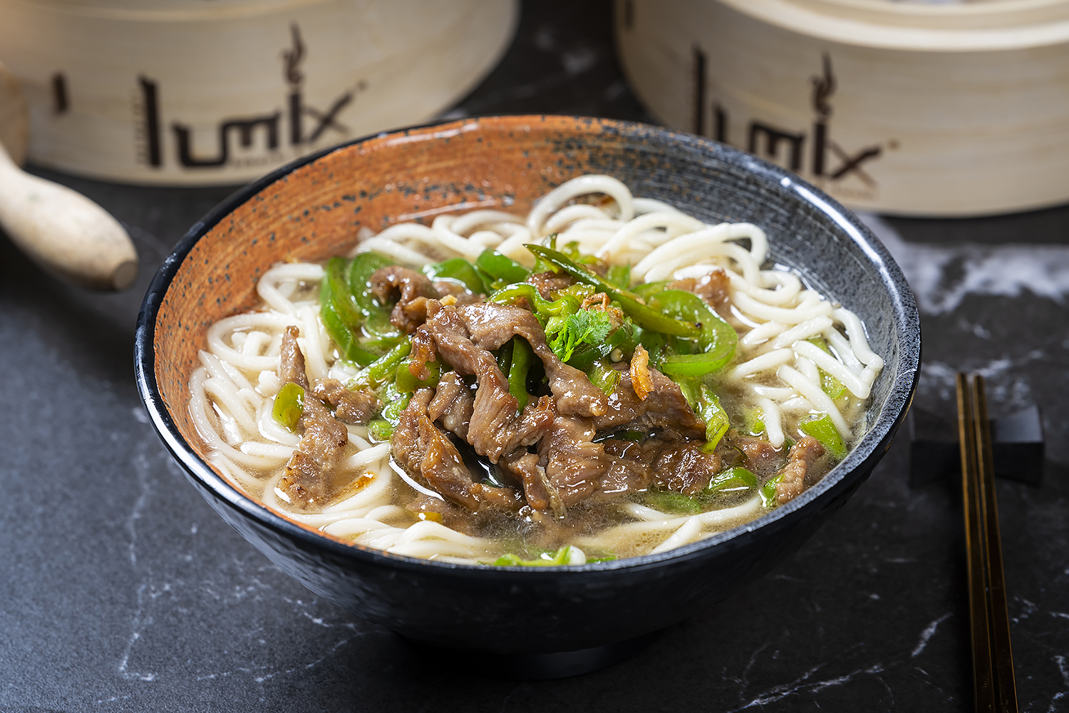 Beef noodle soup with sliced beef, green peppers and cilantro in rustic ceramic bowl. Black marble table, bamboo steamers and chopsticks on side.