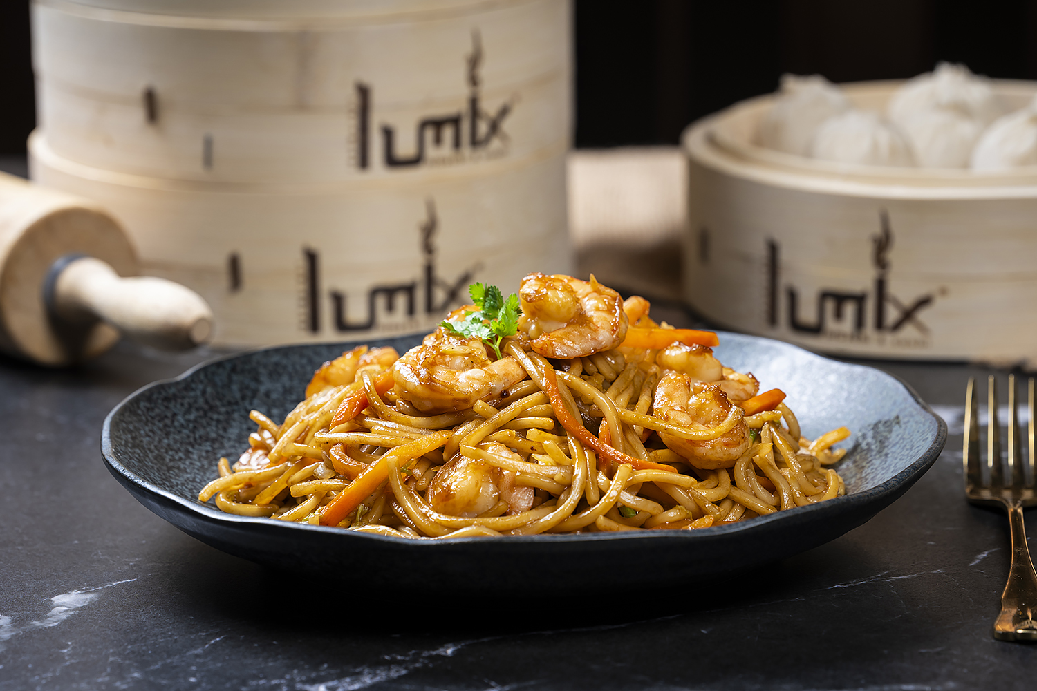 Stir-fried lo mein noodles topped with large shrimp, carrot strips and cilantro. Black textured plate, bamboo steamers and golden fork on marble table.