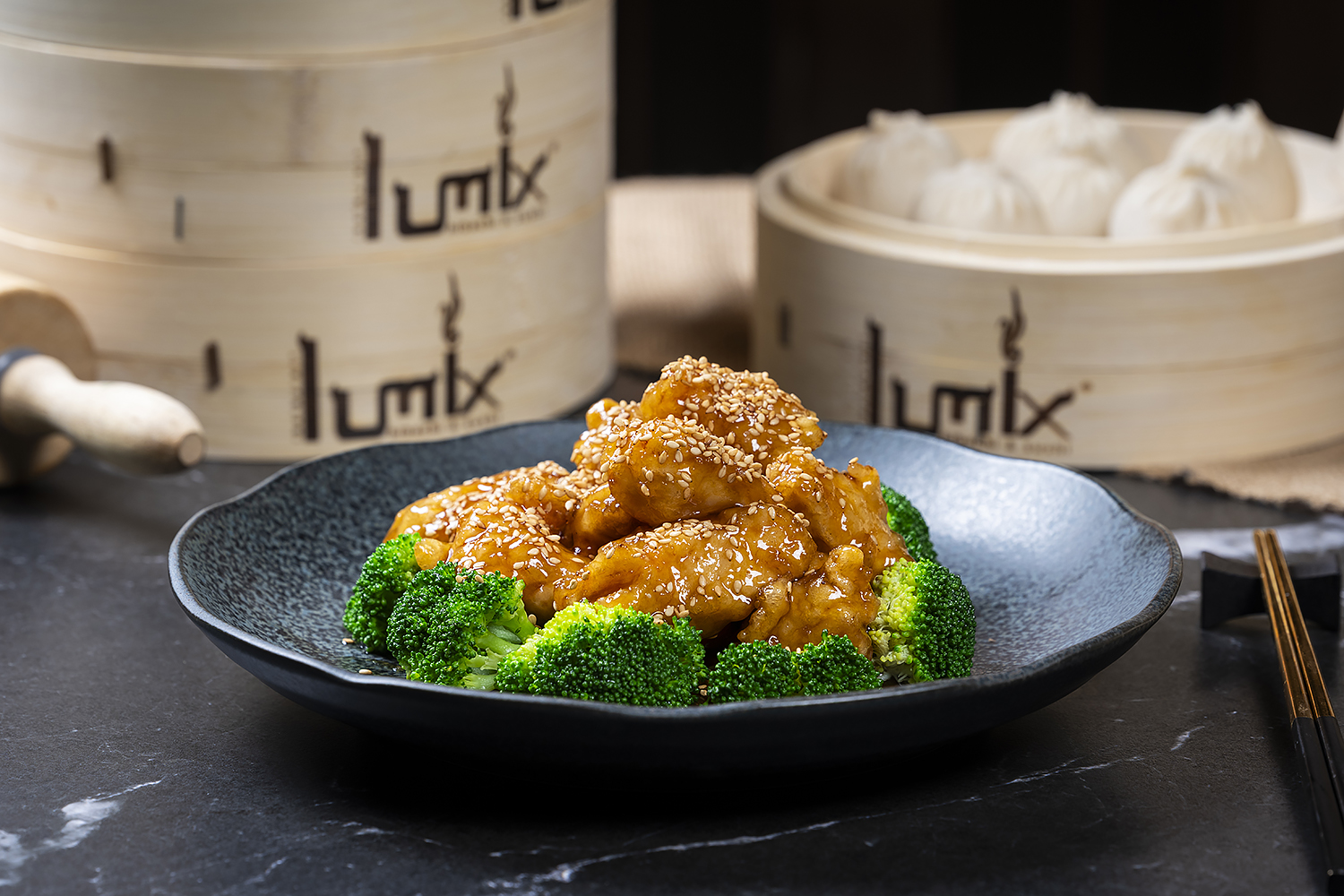 Glazed sesame chicken pieces sprinkled with sesame seeds, served with broccoli. Dark speckled plate, bamboo steamers with dumplings in background.