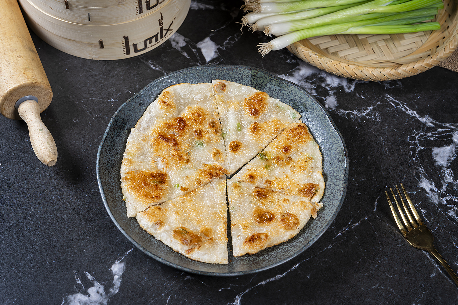 Golden crispy scallion pancake sliced into six pieces on dark plate. Black marble table, rolling pin, fresh green onions and bamboo basket in background.