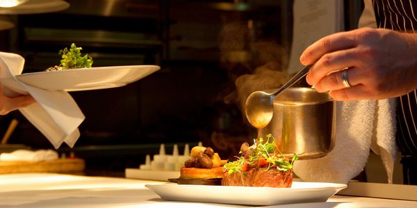 Chef pouring sauce over a gourmet steak garnished with microgreens in a professional restaurant kitchen.