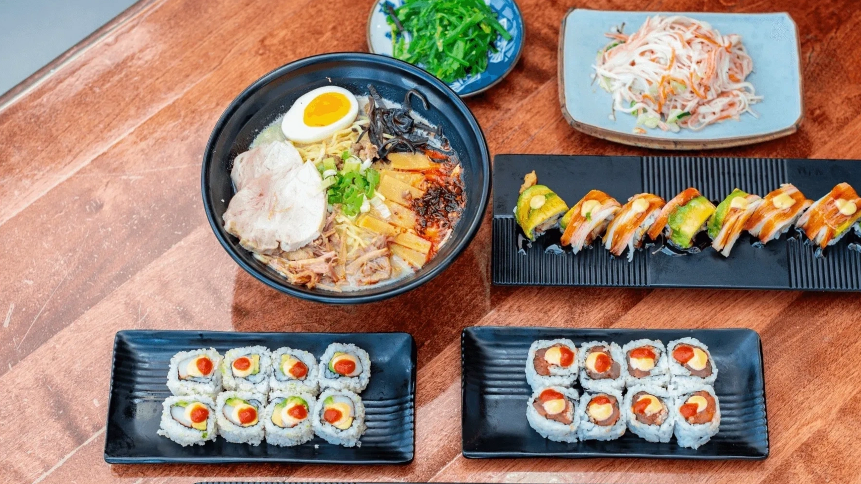 Japanese feast featuring ramen, assorted sushi rolls, seaweed salad, and kani salad on a wooden table.