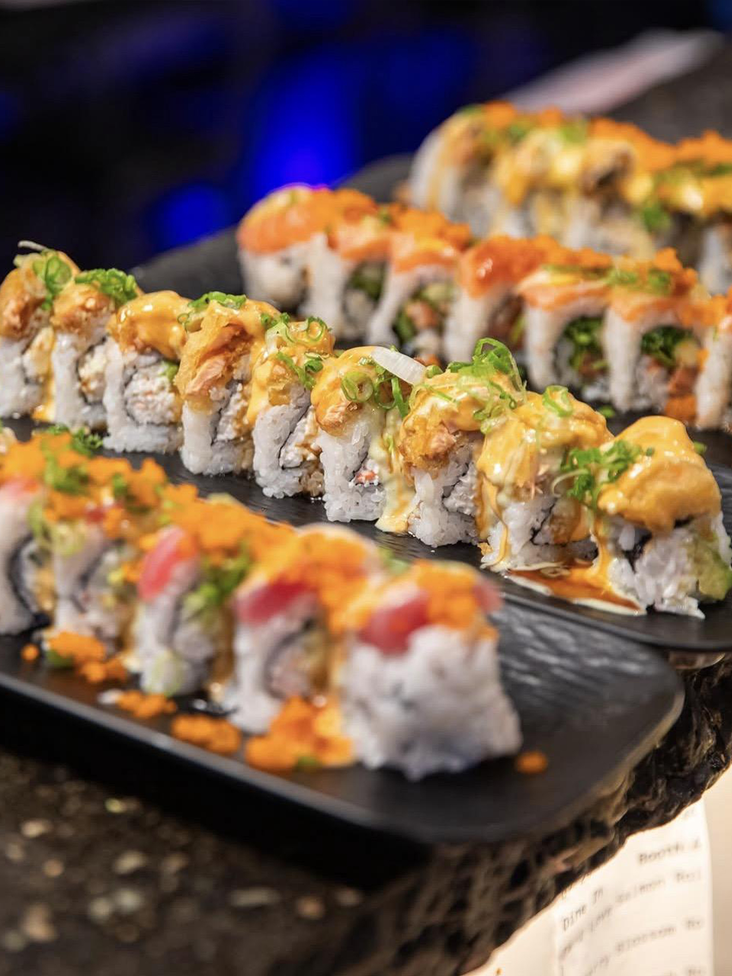 Assorted sushi rolls topped with spicy mayo, green onions, and roe on black plates.