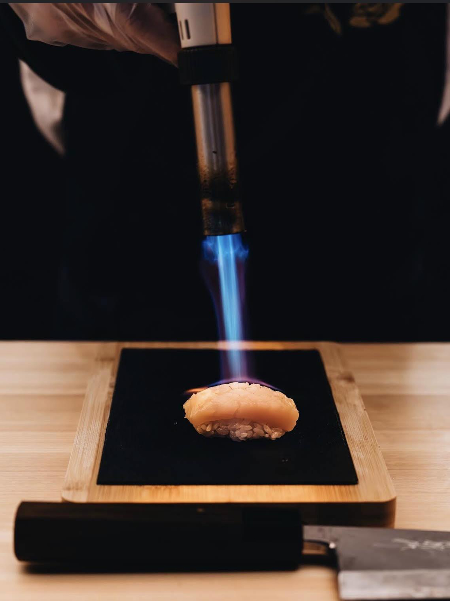 Chef using a blowtorch to sear a piece of nigiri sushi on a wooden board.