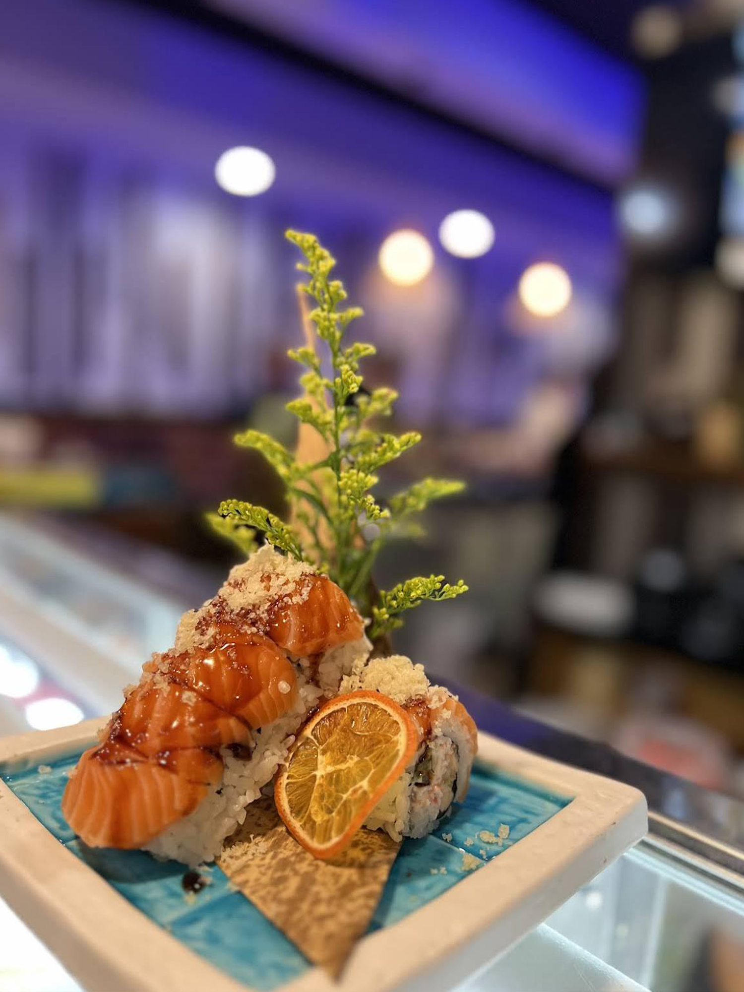 Salmon sushi rolls topped with sauce and crumbs, garnished with a dried orange slice.