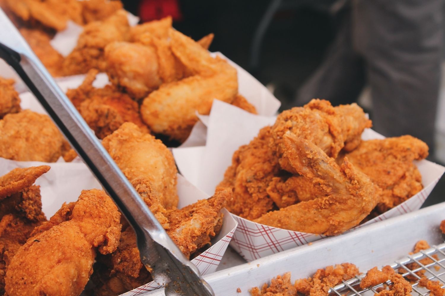 Multiple baskets of crispy fried chicken wings and drumsticks displayed at a food stall with metal tongs in the foreground.