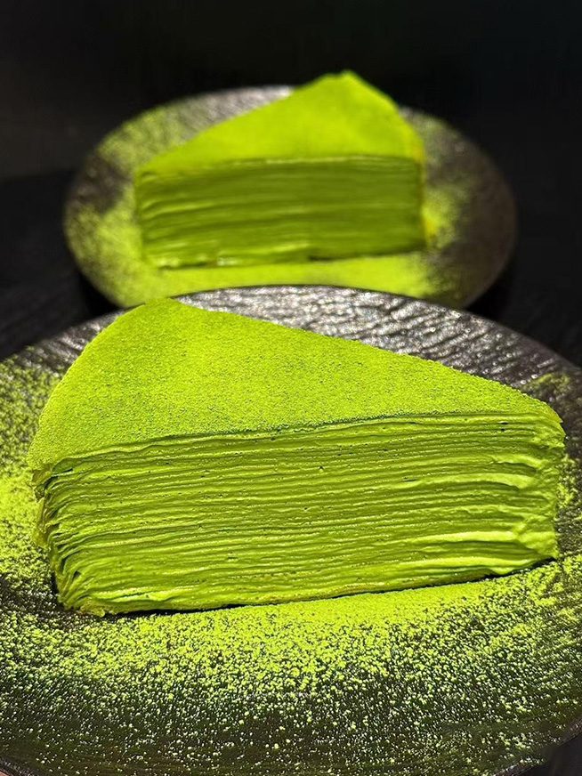 Layered matcha crepe cake dusted with fine green matcha powder on dark textured ceramic plate at Meet Again Cafe, a Cafe in Scotch Plains