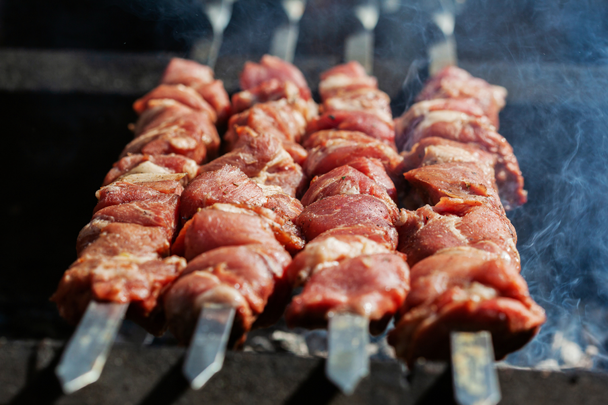 Raw marinated meat chunks threaded onto metal skewers, cooking over an open flame with rising smoke (grill scene)