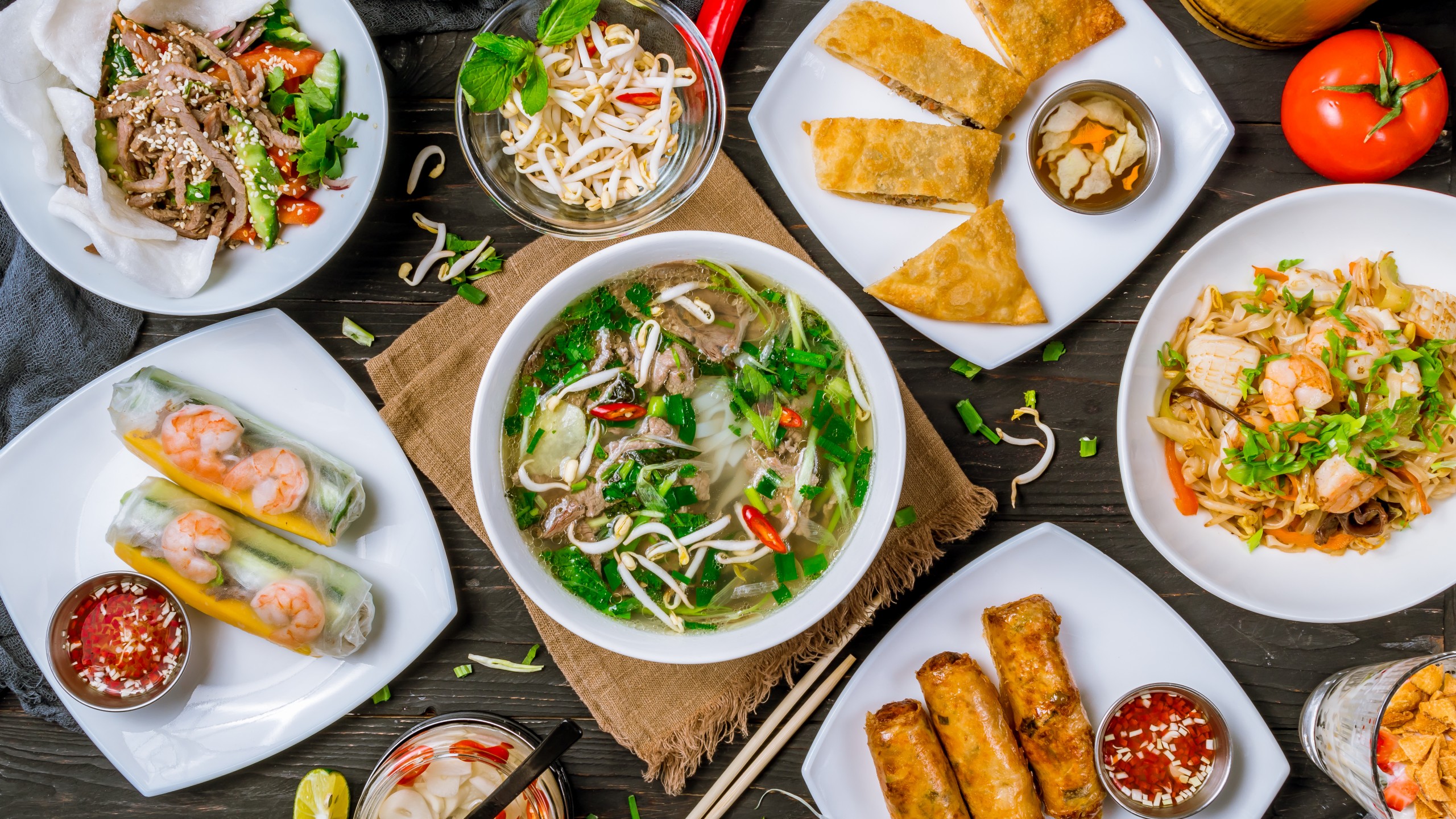 Assorted Vietnamese dishes including pho, spring rolls, fried pastries, stir-fried noodles, and fresh ingredients, arranged on a dark wooden table.