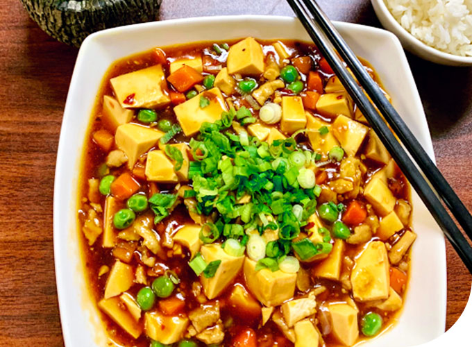 Chopsticks-Chinese-Restaurant-Mapo-Tofu-Kent A plate of Mapo Tofu, with soft tofu cubes in a rich, savory sauce, mixed with peas, diced carrots, and minced meat. Topped with chopped green onions, served with chopsticks, and accompanied by a bowl of white rice on a wooden table at Chopsticks,a Chinese Restaurant in Kent