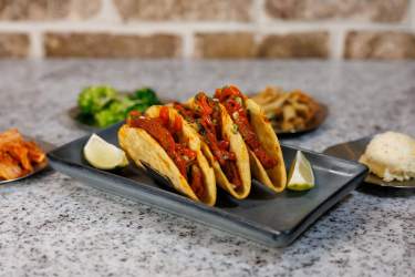 Three tacos with spicy meat, lime wedges, and side dishes on a gray plate
