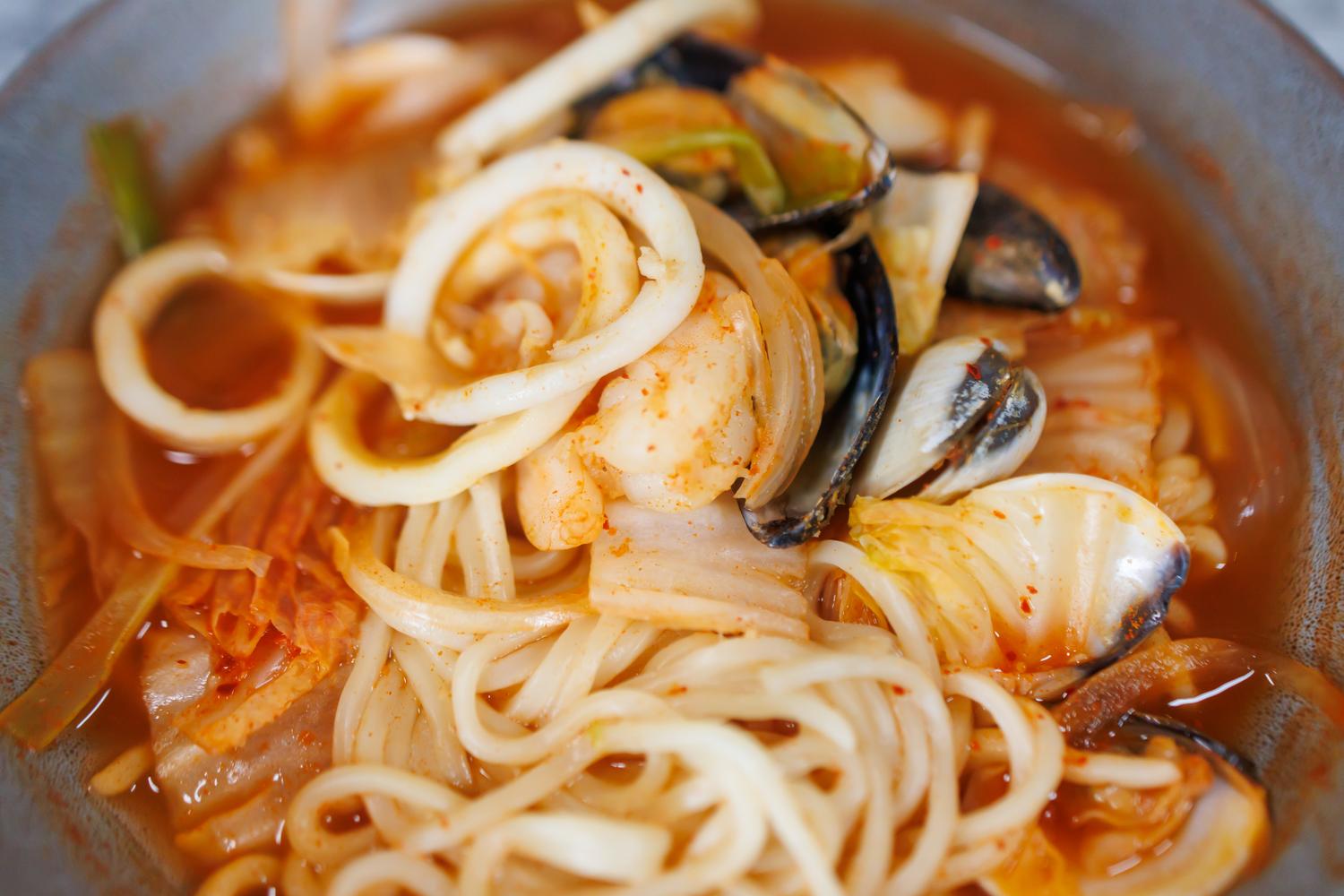 Close-up of spicy Korean seafood noodle soup with mussels, squid, shrimp, onion rings, and cabbage in red broth.