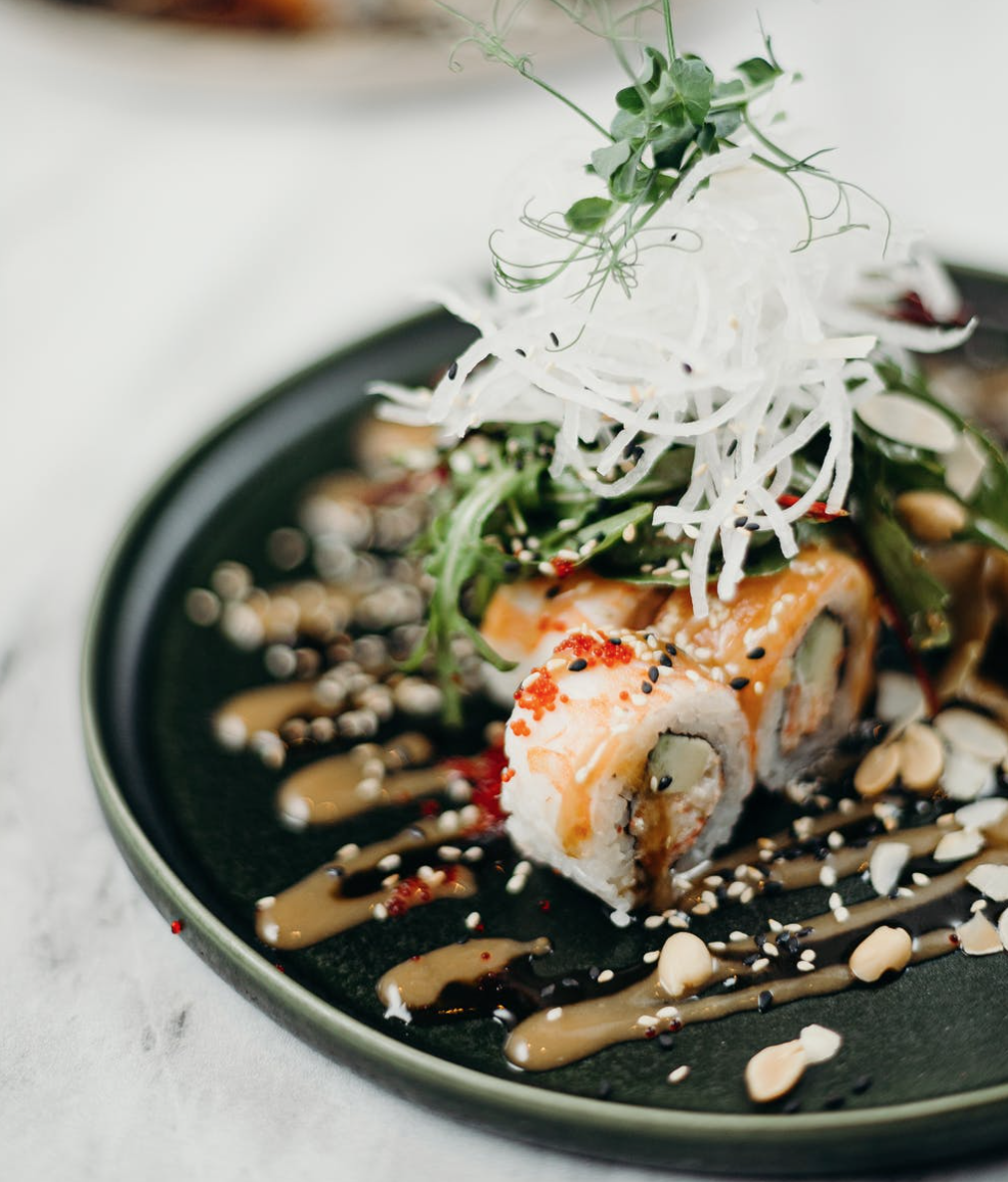 Creative sushi rolls on a black plate, paired with vegetable salad, decorated with microgreens, drizzled with sauce and sprinkled with nut crumbs