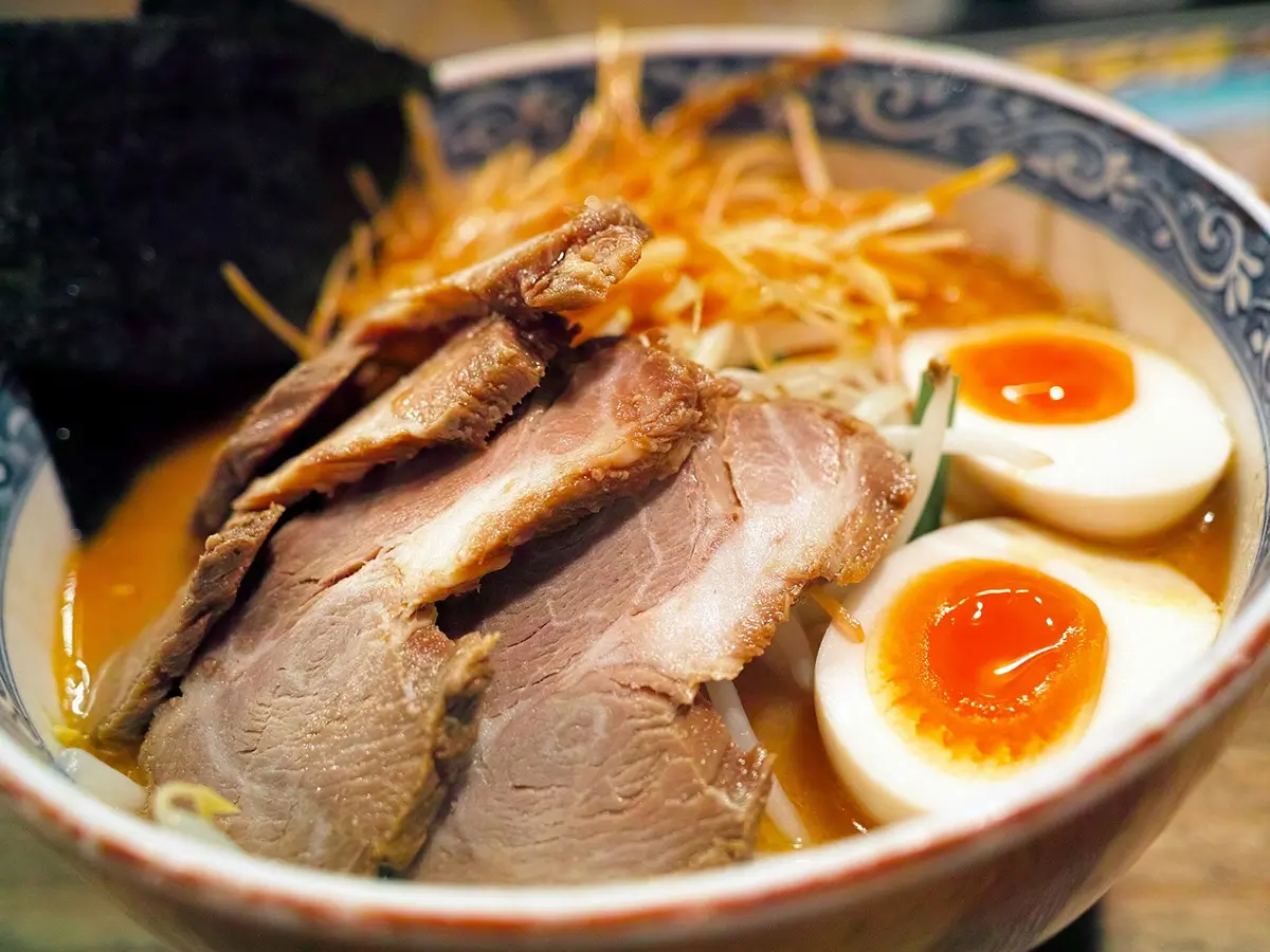 Close‑up of ramen bowl with stacked tender chashu pork slices, two soft‑boiled runny‑yolk eggs, nori seaweed, shredded vegetables, rich orange‑toned broth, ornate patterned ceramic bowl, warm‑toned shallow depth‑of‑field food photograph.